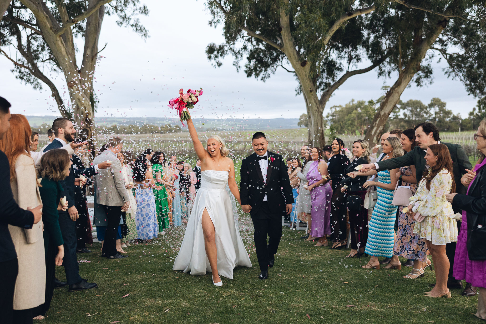 A bride and groom are exiting their ceremony at Mitolo wines. The couple is surrounded by guests throwing petals over them and celebrating. The bride is holding her bouquet high in the air and is mid-cheer.